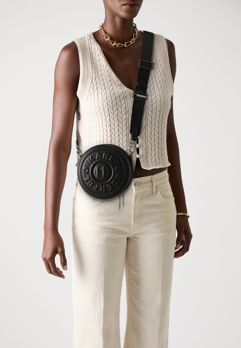 Black circular crossbody bag with embossed branding, worn with a cream knitted vest and cream wide-leg pants, featuring a detachable strap.