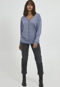 Cardigan blu heather con scollatura a V, maniche lunghe e chiusura con bottoni. Abbinato a jeans scuri con orlo sfilacciato e stivali neri alla caviglia.