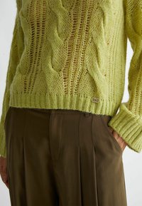 Maglione lavorato a maglia verde lime con motivo a treccia e orlo a coste, abbinato a pantaloni marroni sartoriali con pieghe e tasche laterali.