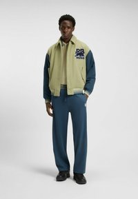 Chaqueta estilo letterman en verde y azul marino con logo, combinada con pantalones de chándal azul marino. Tela suave, ajuste relajado y puños acanalados en contraste. Zapatos negros.