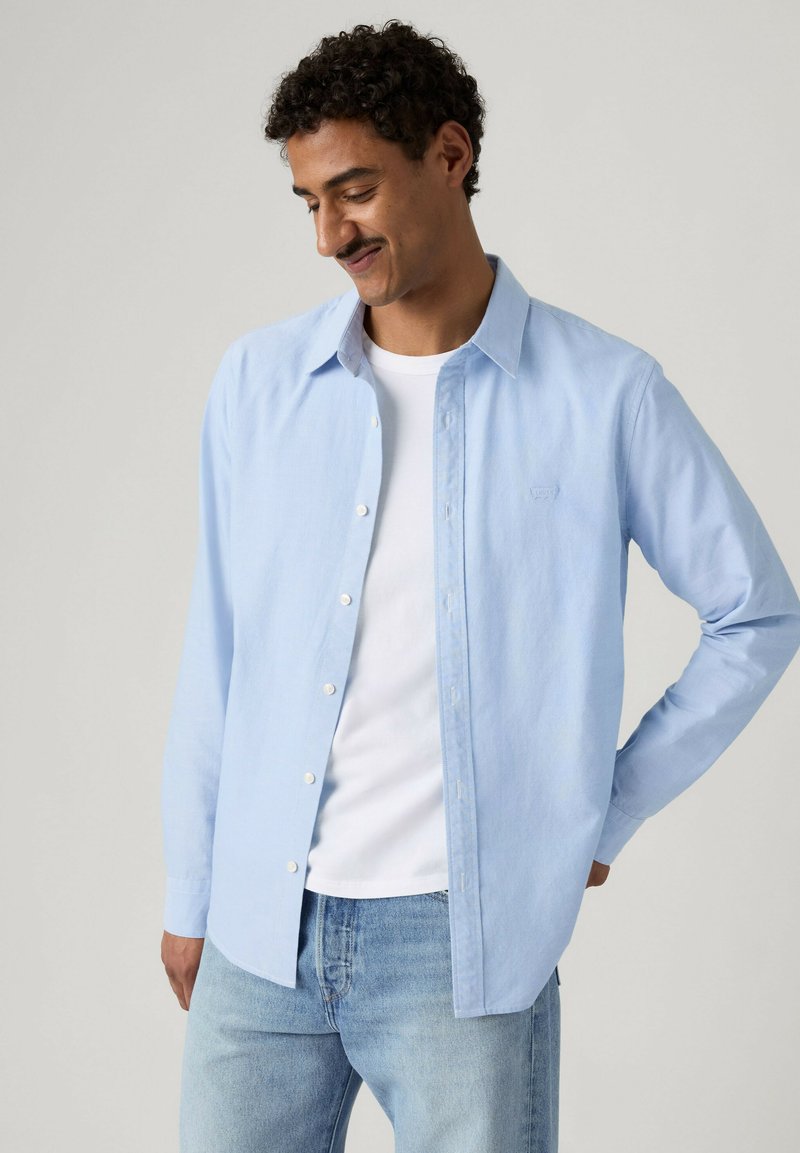 Mand med krøllet hår iført lyseblå skjorte med knapper over en hvid T-shirt og lyseblå jeans, står med hånden på hoften.