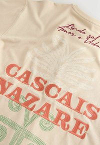 Tissu beige avec texte brodé "CASCAIS NAZARÉ", un motif abstrait de feuille à points blancs, et du texte rouge en cursive en haut.