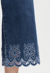 Τελική ραφή φούστας από τζιν με κυματιστή άκρη και κεντημένα λουλουδένια μοτίβα με τελειώματα ματιού σε ανοιχτό μπλε νήμα.