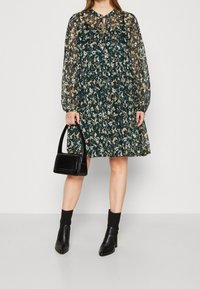 Květinové šaty v tmavě zelené barvě s dlouhými průhlednými rukávy, vrstevnatým designem a výstřihem ve tvaru klíče, doplněné černou strukturovanou kabelkou a kotníkovými botami.