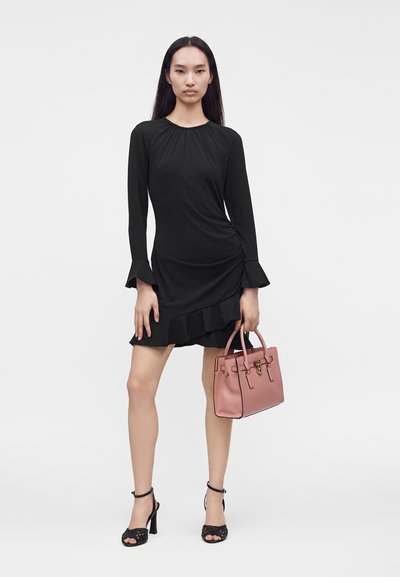 Femme portant une robe noire à manches longues à volants, tenant un sac à main rose et portant des sandales noires à talons hauts sur un fond uni.