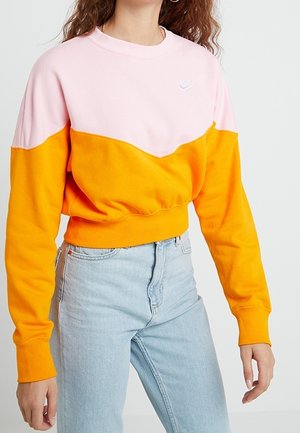 Vrouw draagt een color-block Nike sweatshirt in roze en oranje met lichtblauwe, hooggetailleerde jeans, staand tegen een effen achtergrond.