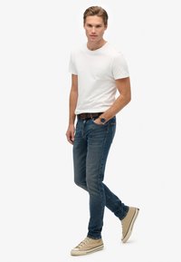 Joven caminando, vistiendo una camiseta de manga corta blanca, jeans azules, zapatillas beige, un cinturón marrón y un reloj de pulsera con esfera azul.
