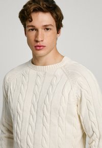 Maglione crema a lavorazione a treccia con scollatura rotonda a costine e motivo testurizzato. Realizzato in un tessuto morbido, presenta maniche lunghe a raglan.