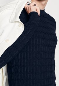 Marineblauwe geribbelde gebreide trui met een hoge hals en een gestructureerd ontwerp. Een witte jas met knopen hangt over één schouder.