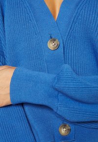 Blå stickad cardigan med V-ringning, ribbad textur och en stor guldknoop. Ärmarna har en figursydd design.