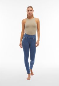 Mujer con una blusa sin mangas beige y vaqueros pitillo azules de talle alto, de pie descalza contra un fondo blanco liso.