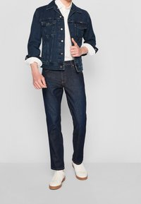 Mörk denimjacka med två bröstfickor, vit skjorta under, mörka denimjeans och vita sneakers med beige sulor.