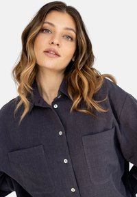 Ciemnoniebieska koszula zapinana na guziki, wykonana z teksturowanej tkaniny, z dwoma kieszeniami na piersiach i srebrnymi guzikami. Kołnierz leży płasko.