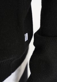 Sort strikket sweater med ribbet tekstur, der har et lille hvidt mærke. Nærbillede viser foldede manchetter og detaljer af stoffet.