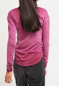 Pitkähihainen, malvanvärinen paita, jossa on teksturoitu, hieman haalistunut viimeistely. Paidassa on pyöristetty helma ja istuvat hihat, yhdistetty tummanharmaisiin housuihin.