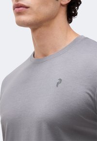 Κοντινό πλάνο ενός άνδρα που φοράει απλό ανοιχτό γκρι μπλουζάκι με στρογγυλή λαιμόκοψη και μικρό, σκούρο γεωμετρικό λογότυπο στο αριστερό στήθος.