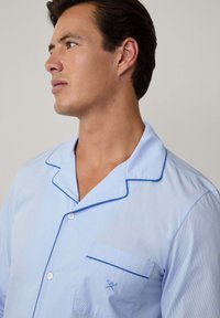 Hellblau gestreifte Schlafanzugbluse mit einem angeschnittenen Kragen, Brusttasche und blauen Paspelakzenten. Hergestellt aus weichem Stoff mit Knopfverschluss.