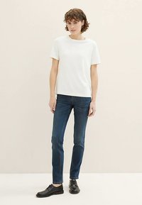 Biała bawełniana koszulka z okrągłym dekoltem, w zestawieniu z ciemnoniebieskimi obcisłymi dżinsami i czarnymi butami. Model stoi na neutralnym tle.