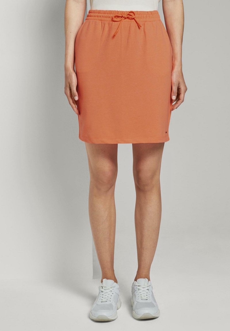 Falda de algodón stretch naranja con cinturilla elástica y cordón. Textura suave, hasta la rodilla, combinada con zapatillas deportivas blancas.