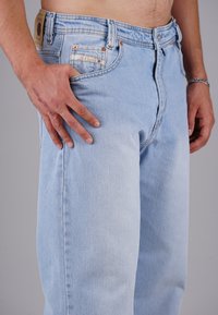 Mann, der hellblaue Jeans trägt, mit einer Hand auf dem Oberschenkel ruhend, zeigt eine Narbe auf dem Handrücken und ein silbernes Armband am Handgelenk.