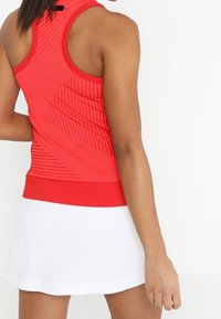 Czerwony top na ramiączkach z teksturowanymi, ukośnymi paskami i jednolitym czerwonym pasem w talii, zestawiony z białą spódnicą. Skupienie na sportowym kroju i materiałach.