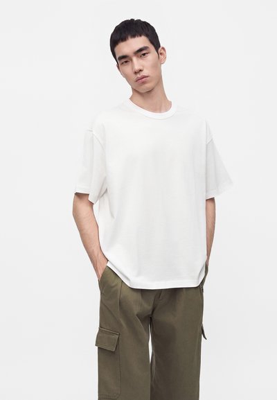 Jeune homme portant un t-shirt blanc à manches courtes oversize et un pantalon cargo vert olive, les mains dans les poches, sur fond uni.