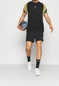 Svart sportt-shirt med gula mönstrade ärmar, tillsammans med svarta shorts och vita fotbollsskor med gröna detaljer. Håller en fotboll.