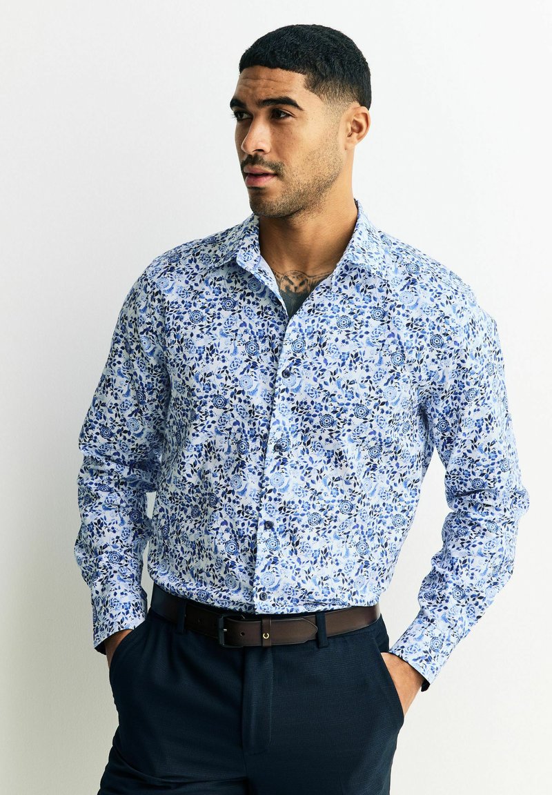 Koszula zapinana na guziki z jasnoniebieskiej tkaniny z kwiatowym wzorem z akcentami w ciemnoniebieskim kolorze. Długie rękawy, kołnierzyk i luźny fason.