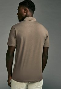Kurzärmeliges, braunes Poloshirt mit kragenförmigem Ausschnitt, glatter Textur und Seitenschlitzen, getragen mit hellen Hosen.