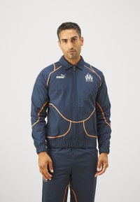 Mornarska športna jakna z oranžnimi akcenti, z zadrgo in rebrastimi robovi. Vključuje logo na prsih in piping dizajn ob ramenih ter straneh.