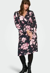 Žena u crnoj haljini dugih rukava s velikim ružičastim cvjetnim uzorkom, crnim tajicama i crnim čizmama do gležnja, stoji s rukama prepletenim.