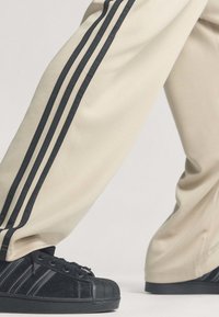 Pantalon beige avec trois bandes noires porté avec des baskets noires présentant un design à trois bandes assorti et des embouts texturés.