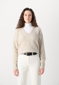 Beige strikket genser med v-hals, teksturert mønster og lange ermer, kombinert med hvite bukser og et svart belte.