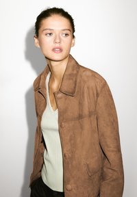 Brun ruskindsjakke med krave og knaplukning, kombineret med en lysegrøn ribbet V-hals top. Baggrunden er helt hvid.