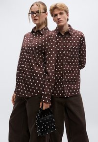 Καφέ πουκάμισα με άσπρες πουά βούλες, με μακριά μανίκια, τσέπη στο στήθος, συνοδευόμενα από σκούρα παντελόνια και μια μικρή μαύρη τσάντα με καρφιά.