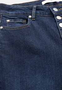 Farkkuhousut, joissa on tumma sininen pesu, korkea vyötärö, näkyvät ompelutiedot sekä metalliset napit edessä.