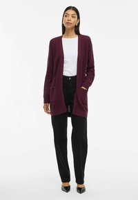 Bordeauxfarbener Strickcardigan mit offenem Vorderteil, langen Ärmeln und zwei vorderen Taschen, kombiniert mit einem weißen Oberteil und schwarzen Hosen. Schwarze spitze Schuhe.