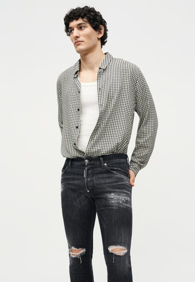 Νέος άνδρας με κατσαρά μαλλιά φοράει μαύρο-άσπρο καρό πουκάμισο με κουμπιά πάνω από λευκό φανελάκι και μαύρα φθαρμένα τζιν με σκισμένα γόνατα.