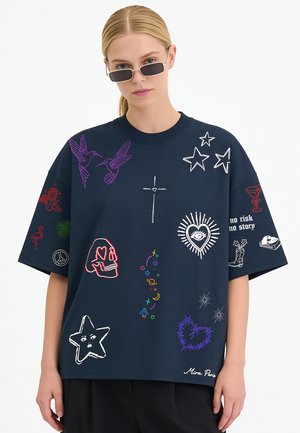 Jeune femme aux cheveux blonds et lunettes de soleil porte un t-shirt oversize bleu marine décoré de symboles graphiques colorés et de texte.