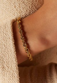 Gouden gedraaide armband rustend op de huid, met een gevlochten ontwerp en een gladde textuur tegen een lichte, gestructureerde stoffen achtergrond.