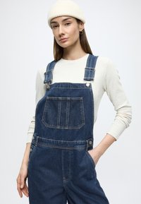 Tumedat sinist tooni teksast kombinesoon reguleeritavate rihmade, esitasku ja külgsete nööpidega. Kandmiseks heledavärvilise soonikkoega pika varrukaga särgi ja mütsiga.