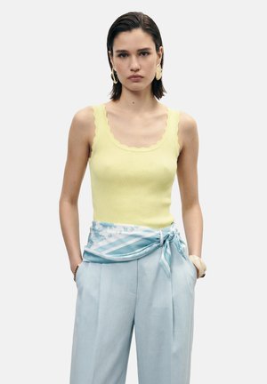 Giovane donna che indossa un top giallo con bordo smerlato, pantaloni azzurri a vita alta con cintura a foulard annodata e grandi orecchini rotondi.