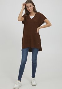 Pull en tricot marron oversize avec un col en V, des manches courtes et des fentes sur les côtés. Associé à un jean skinny bleu et des baskets blanches.