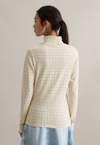 Lange mouwen strakke top in lichtgeel met een ruitpatroon in bruin. Voorzien van een hoge halslijn en een gestructureerde stof, met een rechte zoom.