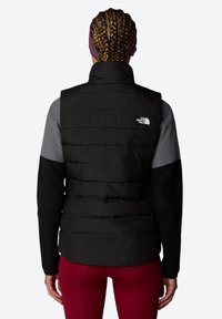 Svart puffervest med quiltet design, høy krage og logo på øvre rygg. Langermet grå og svart skjorte under, kombinert med røde leggins.