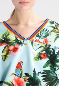 Halenka s akváriem inspirovaným motivem, na světlomodrém pozadí s živými květinovými a papouščími vzory, doplněná vícebarevným výstřihem do V.