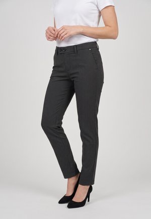 Donna con pantaloni slim fit a righe scure, camicia bianca a maniche corte e scarpe nere con tacco alto, in piedi davanti a uno sfondo semplice.