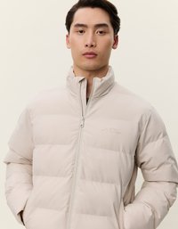 Casaco puffer em bege claro com padrão de cosido horizontal, fecho de zíper total, gola alta e detalhe de logotipo discreto no peito.