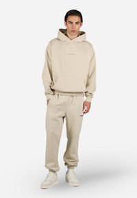 Beige genser med frontlomme og hette, kombinert med matchende joggebukser. Begge har en subtil logo. Glatt tekstur, avslappet passform.
