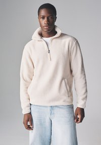 Bluza polarowa z suwakiem do połowy długości w kolorze kremowym, o miękkiej fakturze, z boczną kieszenią i kontrastowym zamkiem. Połączona z jasnoniebieskimi dżinsami.